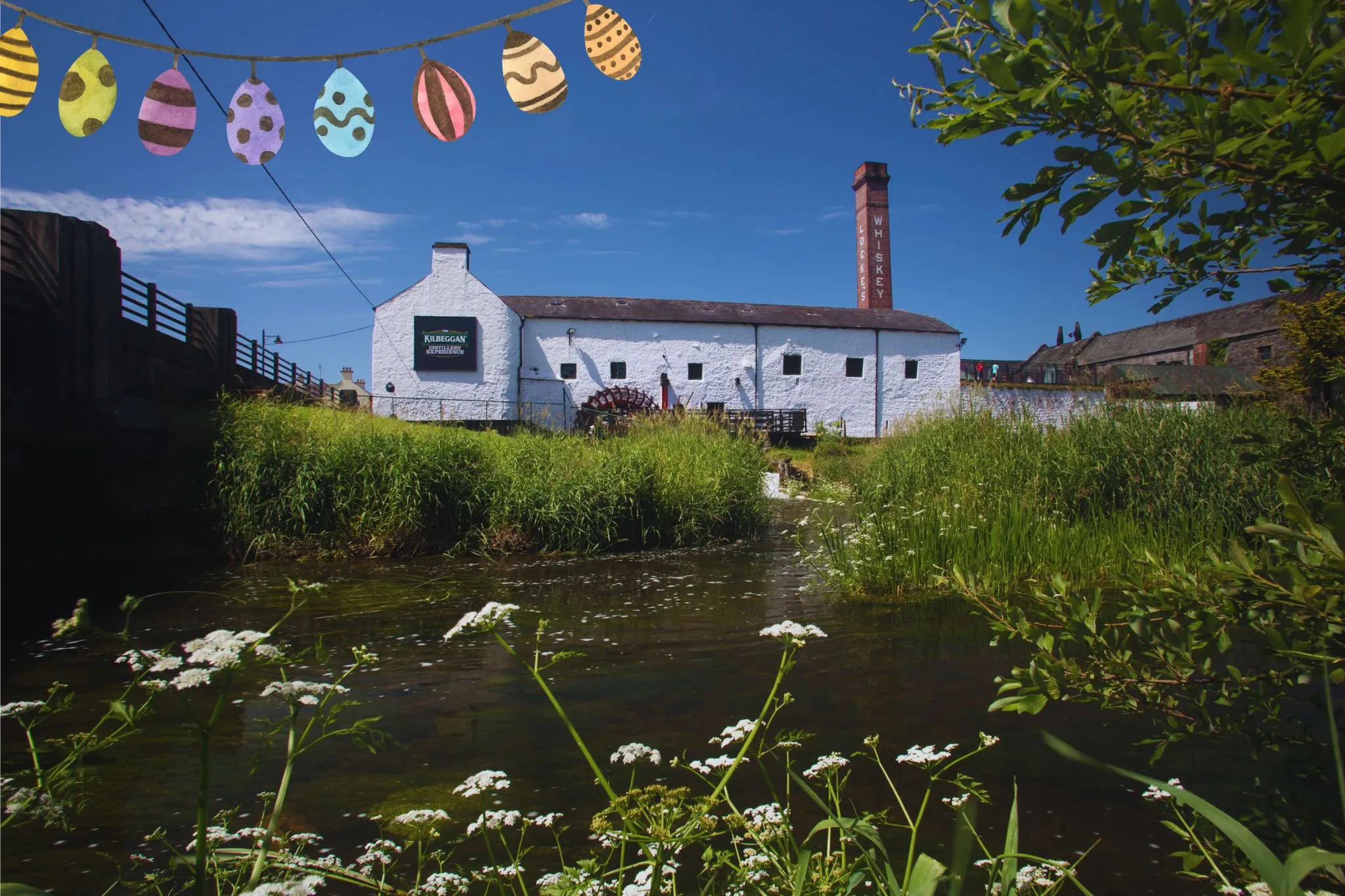 Scenic view of a white whiskey distillery by a stream, adorned with colorful Easter egg decorations against a clear blue sky.